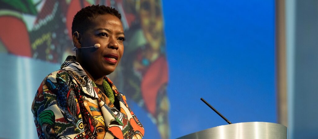 A woman wearing a colorful patterned jacket speaks at a podium with a microphone headset, against a blue background with a large, vibrant image or pattern partially visible behind her.