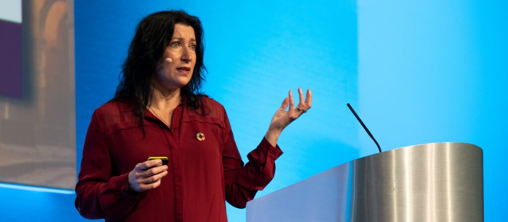 A woman with long dark hair, wearing a red shirt and headset microphone, stands beside a podium, gesturing with her hand while speaking in front of a blue background.