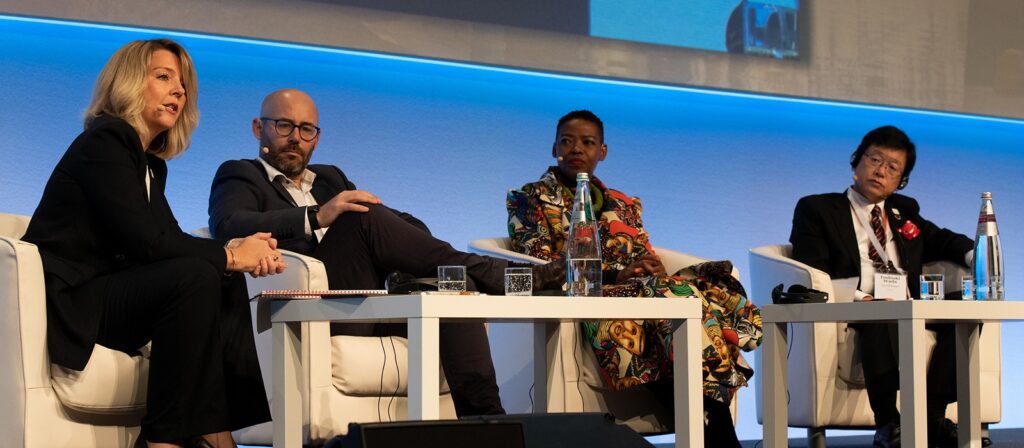 Four people sit on stage in white chairs, engaged in a panel discussion. Each has a microphone and water bottles are on the table in front of them. The background is blue and white with a large screen above.