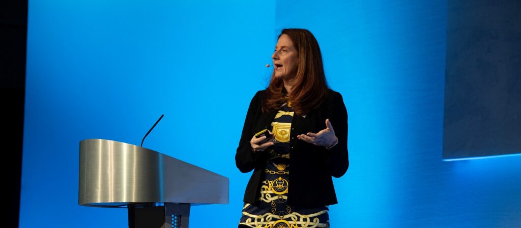 A woman with long brown hair, wearing a blazer and a patterned dress, speaks on stage with a headset microphone, standing beside a modern podium against a blue background.