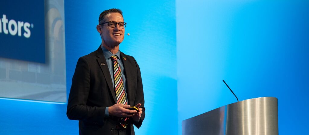 A man in a suit with a colorful striped tie speaks on stage at a podium, smiling and wearing a headset microphone, with a blue background behind him.