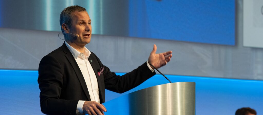 A man in a suit and headset microphone stands at a podium, gesturing with his hand while speaking at a conference or event, with a blue background behind him.