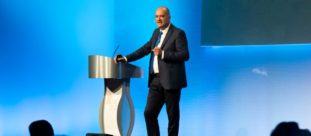 A man in a suit stands on a stage next to a modern podium, speaking into a headset microphone. The background is blue, and an audience is partially visible in the foreground.