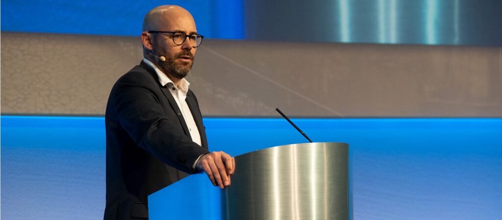 A man wearing glasses and a suit speaks at a podium with a microphone headset in a modern, blue-lit conference setting.