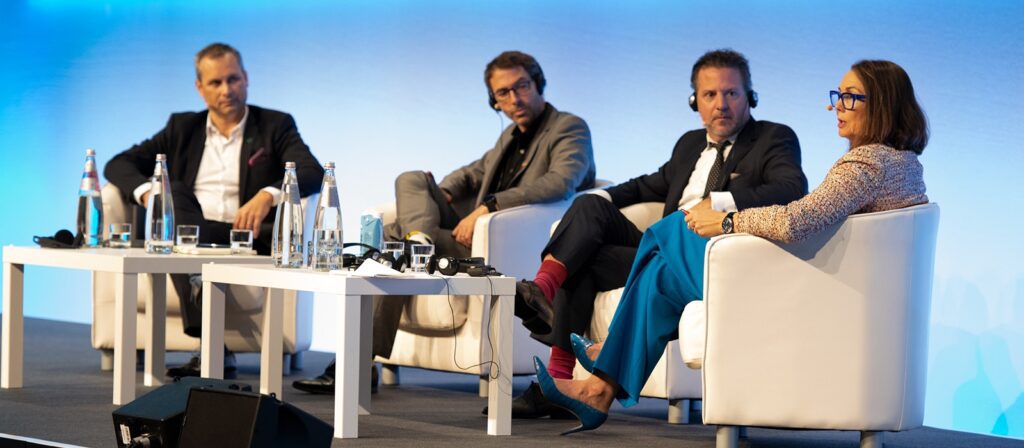 Four people sit on white chairs on a stage, engaged in a panel discussion. Three men wear suits, and one woman in blue pants and glasses speaks while the others listen. Tables with water bottles and microphones are in front of them.
