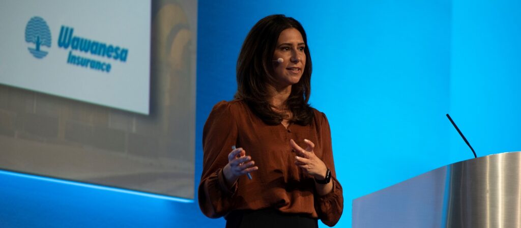 A woman wearing a brown blouse speaks on stage at a podium with a wireless headset microphone. Behind her is a screen displaying the Wawanesa Insurance logo on a blue background.