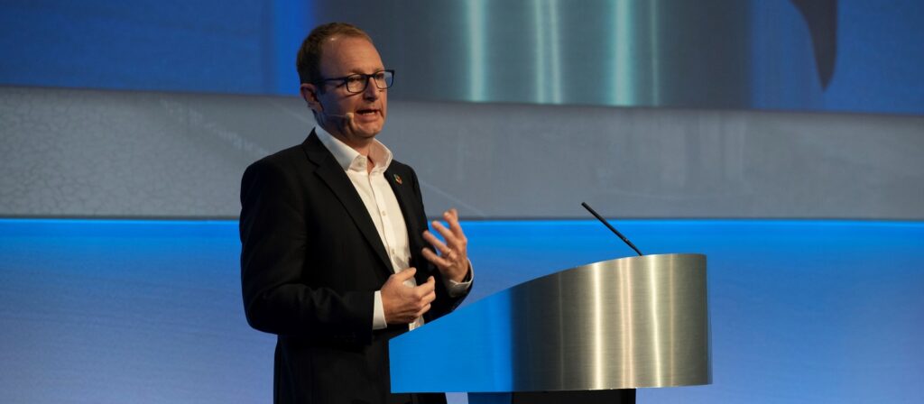 A man wearing glasses and a suit speaks into a headset microphone while standing behind a curved podium on a stage with blue lighting.