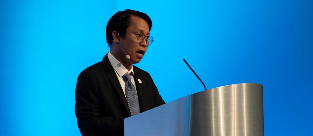 A man in a suit and tie, wearing glasses and a headset microphone, speaks at a metallic podium against a blue background.