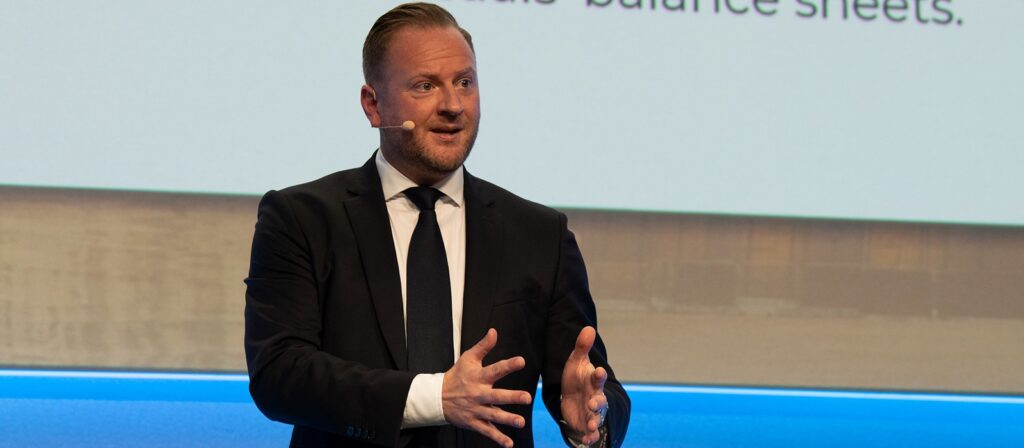 A man in a suit and tie speaks on stage with a headset microphone, gesturing with his hands. A projection screen with text is visible behind him.