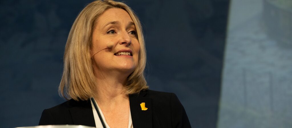 A woman with blonde hair wearing a black blazer and a headset microphone is speaking on stage. She has a yellow pin on her lapel and is smiling, with a blue background behind her.
