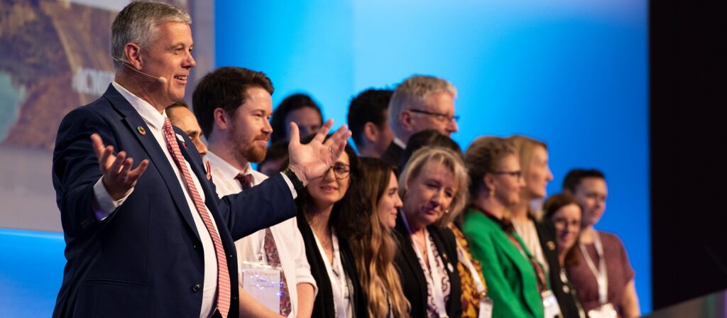 A man in a suit gestures while speaking on stage to an audience; a group of people stand beside him in a row, all facing forward and smiling, with a blue background behind them.