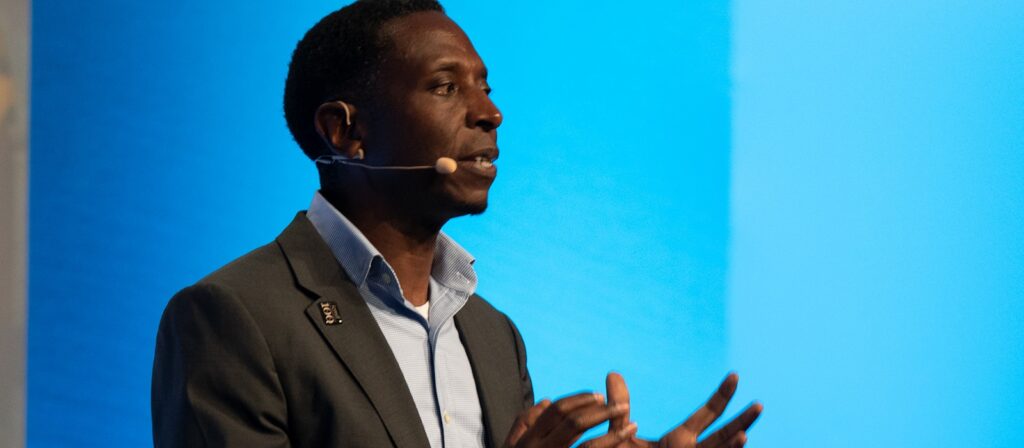 A man in a suit speaks on stage wearing a headset microphone, gesturing with his hands against a blue background.