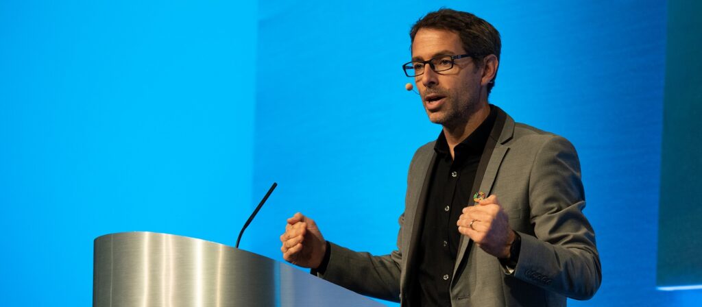 A man wearing glasses and a gray blazer speaks passionately at a podium with a microphone headset, gesturing with both hands against a blue background.