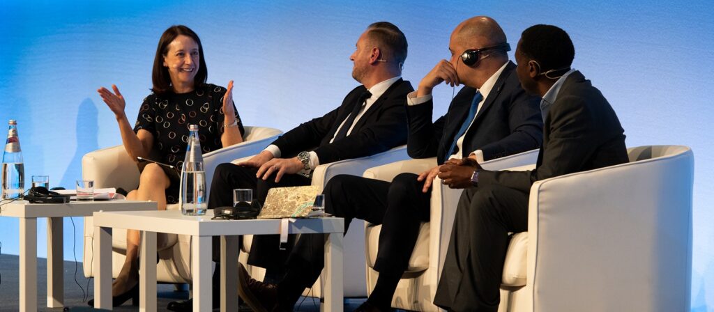 Four people in business attire sit on stage in white chairs, engaged in a lively panel discussion. One woman is gesturing while speaking; three men listen attentively. Bottles of water and microphones are on the table.
