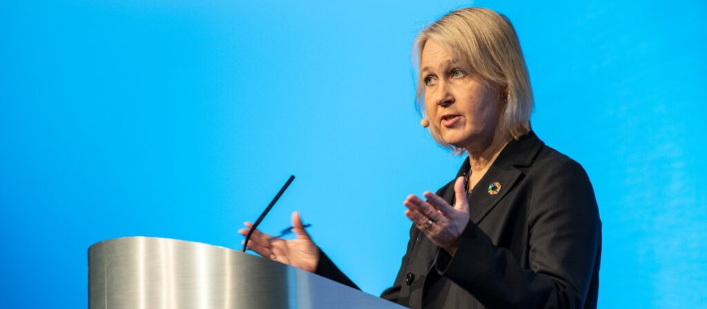 A woman in a black suit speaks at a podium with a microphone headset. She gestures with her hands against a blue background.