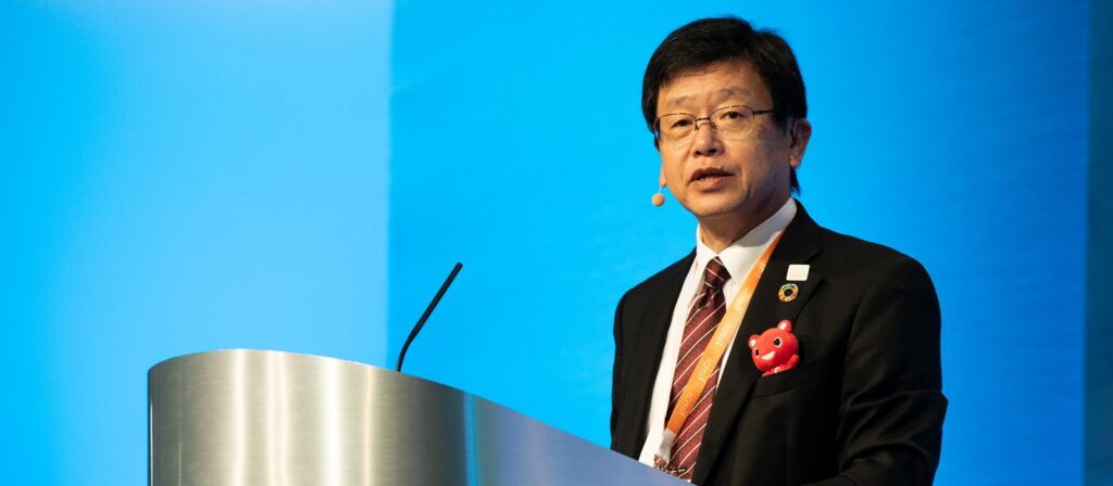 A man in a suit and tie stands at a podium, speaking into a microphone headset, with a blue background behind him. He has a red decorative pin on his jacket.