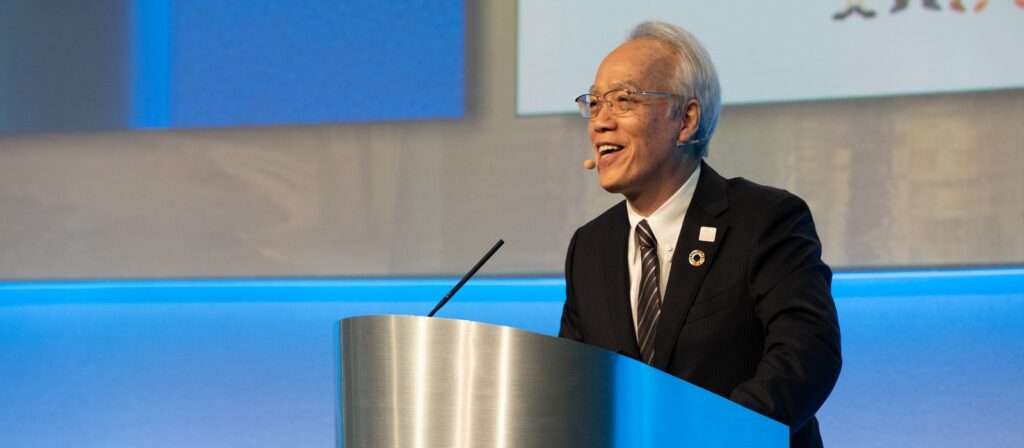 An older man in a suit and tie speaks at a podium with a microphone headset. He is smiling, and there is a blue and white background behind him with some text and graphics partially visible.