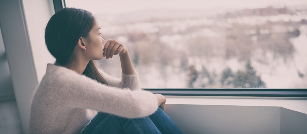 A woman sits by a large window, resting her chin on her hand, and looks out at a snowy landscape. She appears thoughtful, wearing a light sweater and jeans with her hair down.