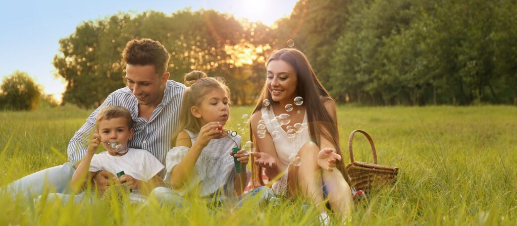 A family of four sits on grass in a sunlit field, smiling and blowing bubbles together. A picnic basket is nearby, and trees are visible in the background.