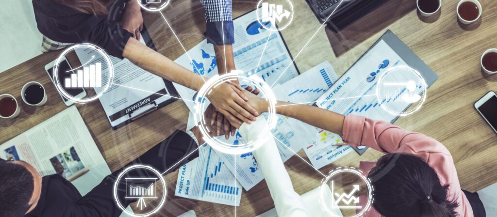 Five people put their hands together over a table covered with business charts and documents, symbolizing teamwork and collaboration, with digital icons showing data and growth graphics overlaid on the image.