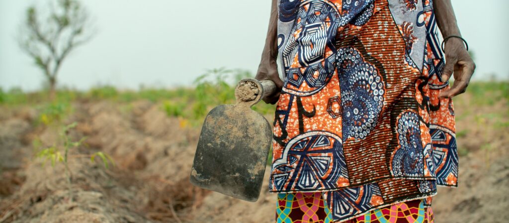 A person wearing a colorful patterned skirt stands in a field, holding a muddy hoe with one hand. The background shows dry soil, sparse plants, and a tree. The persons face is not visible.
