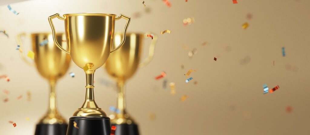 Three golden trophies stand side by side with colorful confetti falling around them, set against a light beige background, symbolizing celebration and achievement.