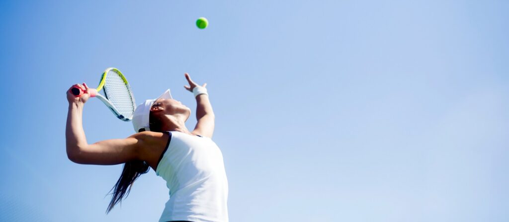 A female tennis player in a white outfit prepares to serve, tossing a tennis ball into the air with her racket ready, against a clear blue sky.