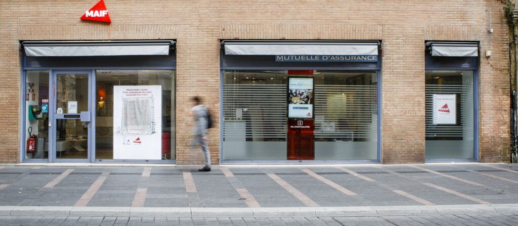 A person walks past the glass storefront of a MAIF insurance office, labeled Mutuelle d’Assurance, with posters and signage visible in the windows on a paved city sidewalk.