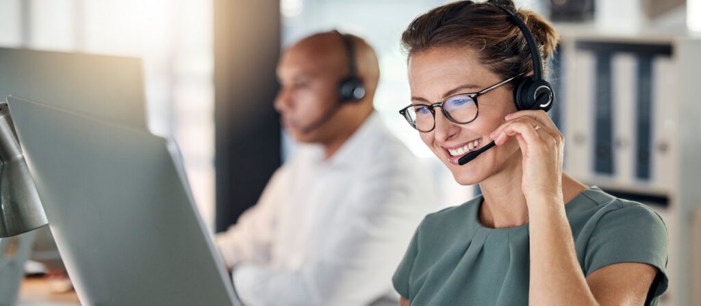 A woman wearing glasses and a headset smiles while working at a computer in an office. In the background, a man wearing a headset also works at a computer.