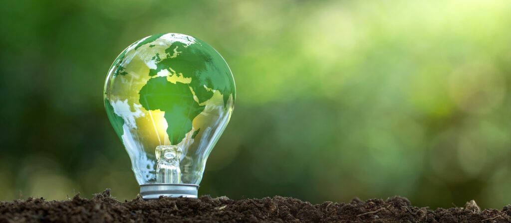 A light bulb with an image of Earth on its surface sits on soil, symbolizing eco-friendly energy or environmental sustainability, with a blurred green nature background.
