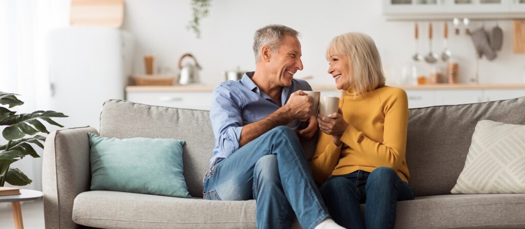 An older man and woman sit close together on a couch, smiling and holding mugs. The living room is bright and cozy, with light-colored decor and kitchen items visible in the background.