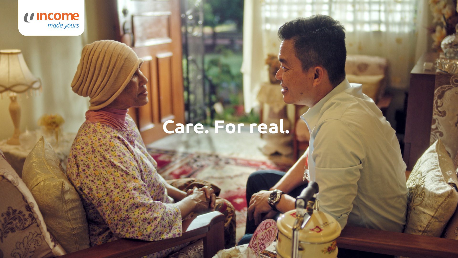 A man gently holds hands and smiles at a woman wearing a headscarf, sitting in a cozy room. The words Care. For real. appear in the center. The Income logo is in the upper left corner.