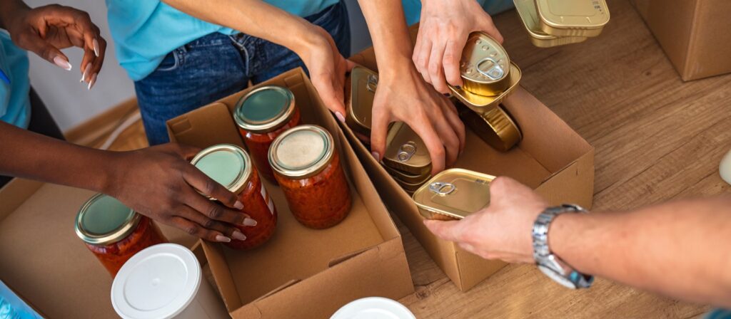Several people pack jars of food and canned goods into cardboard boxes on a wooden table, working together as part of a food donation or volunteer activity.