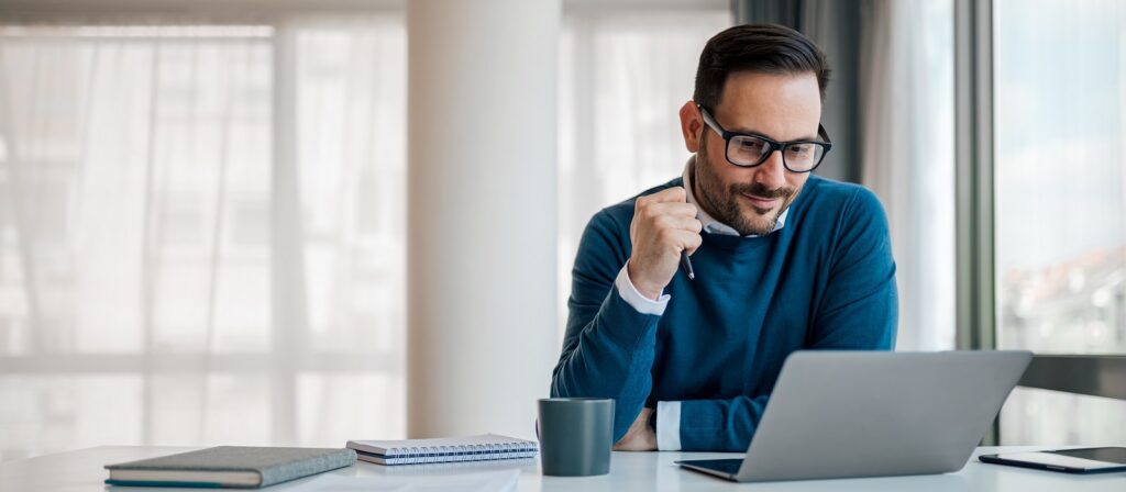 A man wearing glasses and a blue sweater smiles while looking at his laptop. He is sitting at a desk with a notebook, pen, and coffee mug in a bright, modern office with large windows.