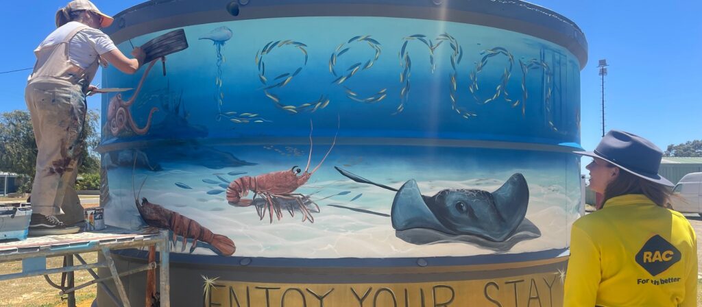 Two people work on painting a mural featuring marine life, including a stingray and prawns, on a large cylindrical tank. The word Carnarvon is visible on the mural. One person stands on scaffolding, the other observes.