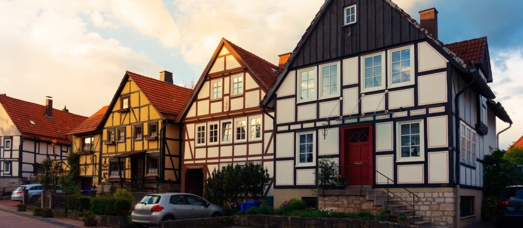 A row of traditional half-timbered houses with red-tiled roofs, large windows, and parked cars in front, basking in warm sunlight on a quiet street.