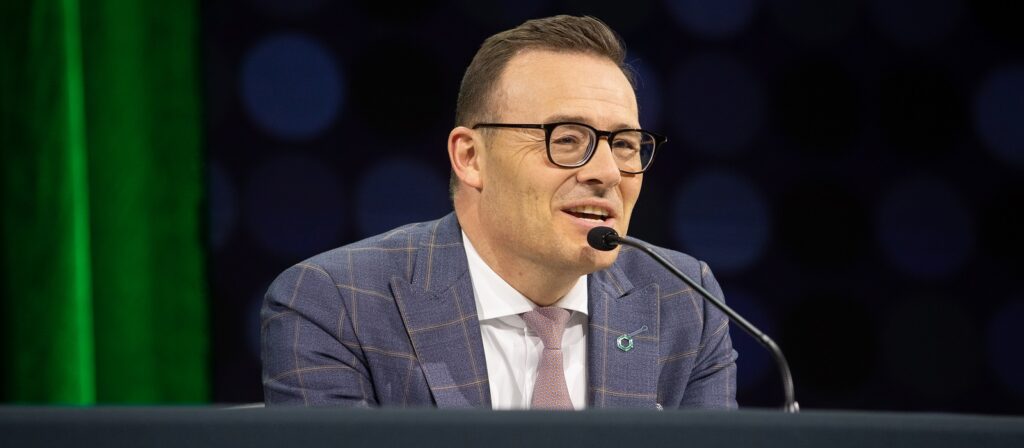 A man in a plaid suit and glasses speaks into a microphone at a table, with a dark background and a green vertical light behind him.