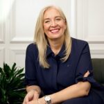 A smiling woman with long blonde hair, wearing a navy blue blouse, sits on a chair next to a green plant in a bright room with white paneled walls.