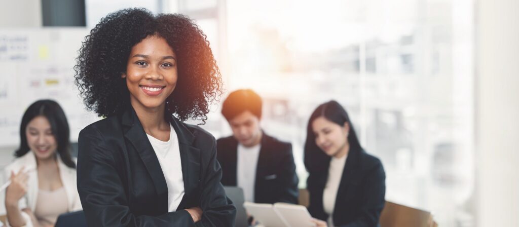 A confident woman with curly hair and a black blazer stands smiling in the foreground, while three colleagues work together on laptops and tablets in the blurred background of a bright office.