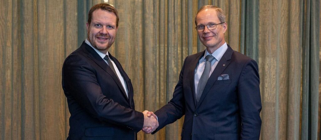 Two men in suits are shaking hands and smiling at the camera, standing in front of beige curtains.