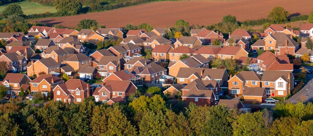 Aerial view of a suburban neighborhood with rows of similar detached houses featuring red or brown roofs, surrounded by trees and open fields in the background.