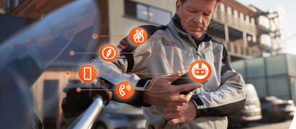 A man in a motorcycle jacket interacts with a smart device on his sleeve, with digital icons including a phone, pen, peace sign, tape, and headset overlaid to illustrate smart wearable technology features.
