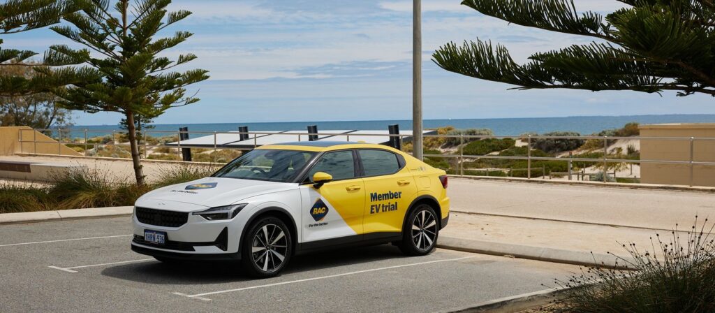 A white and yellow electric car with Member EV trial written on the side is parked near a beach, with sand dunes, ocean, and trees visible in the background under a partly cloudy sky.
