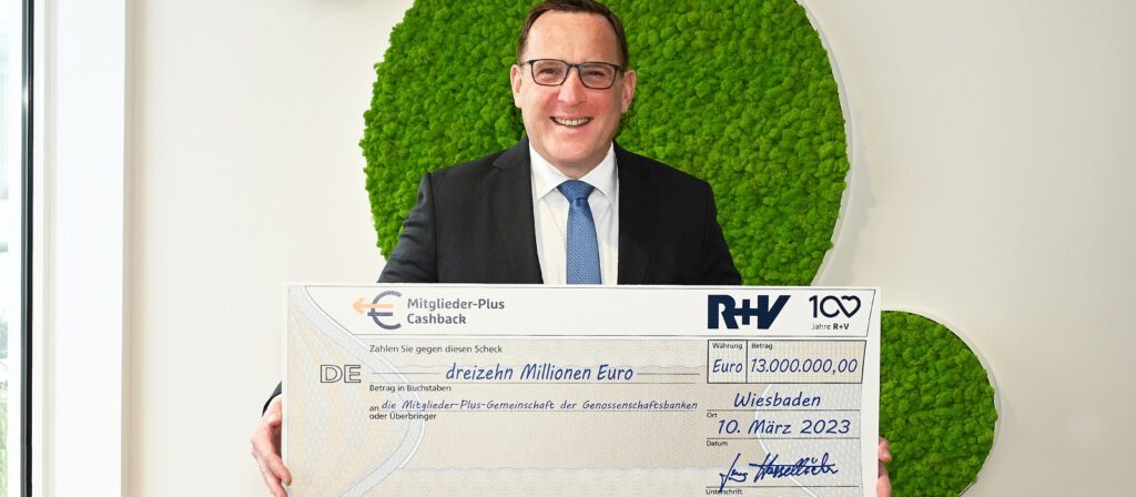 A smiling man in a suit and glasses holds a large ceremonial check for 13 million euros, dated March 10, 2023, in Wiesbaden, with bank and organizational logos visible.