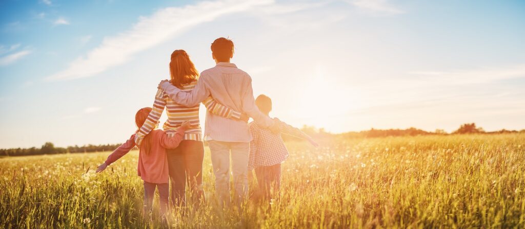 A family of four, standing in a sunlit field, faces the horizon. Parents have their arms around each other, while the children stand on either side with outstretched arms. The sky is bright and clear.