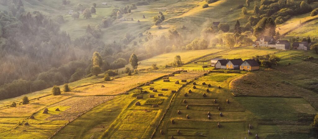 A scenic rural landscape at sunrise, featuring rolling green hills, scattered trees, haystacks, and a few houses with sunlight casting warm golden tones across the fields.