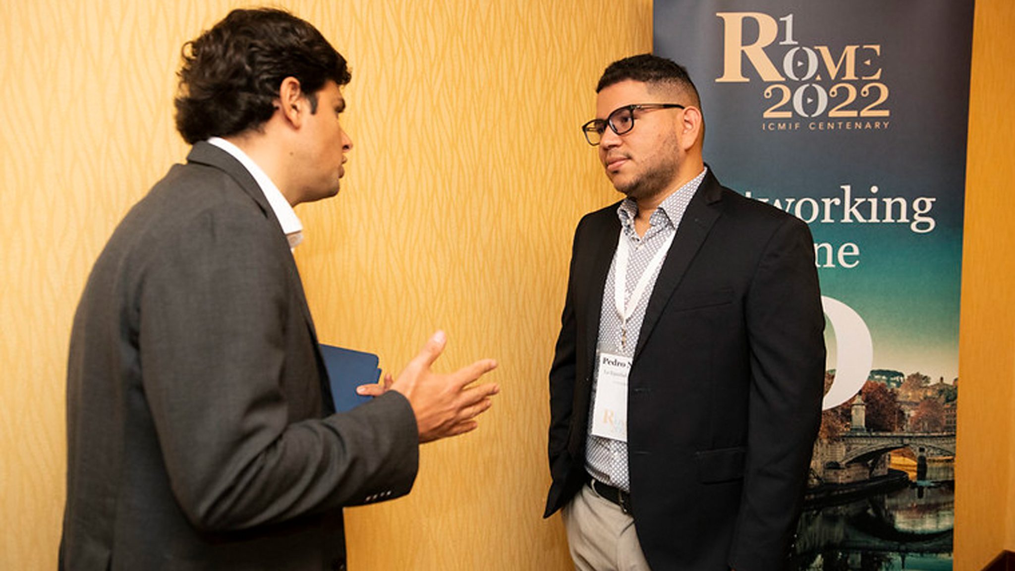 Two men in business attire have a conversation in front of a “Rome 2022” conference banner against a yellow wall, suggesting a professional networking event.