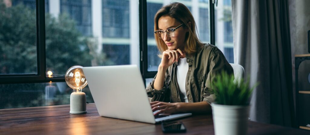 A woman wearing glasses works on a laptop at a wooden desk near a window, with a glowing lamp, plant, and smartphone beside her.