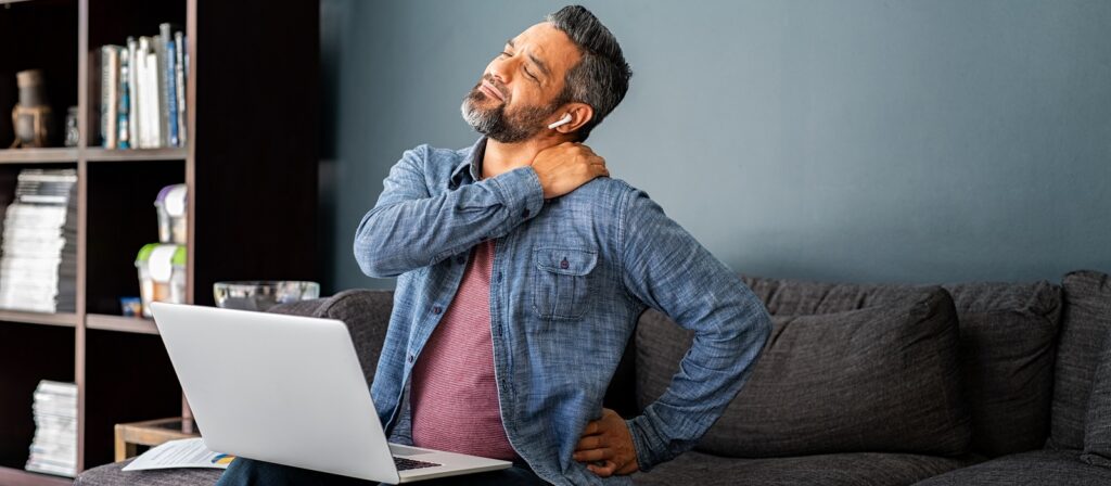A man sitting on a couch in front of a laptop, wearing headphones and holding his neck in discomfort, appearing to have neck or back pain.