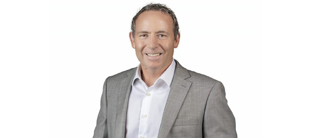 A man with short curly hair wearing a light gray suit jacket over a white dress shirt, smiling, standing against a plain white background.